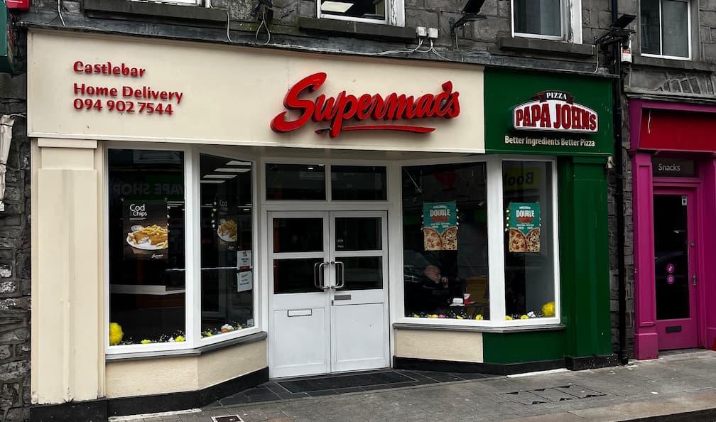 Supermac's and Papa John's restaurant exterior in Castlebar, Ireland. Features dual branding on a traditional stone building, beige Supermac's front, green Papa John's front, 