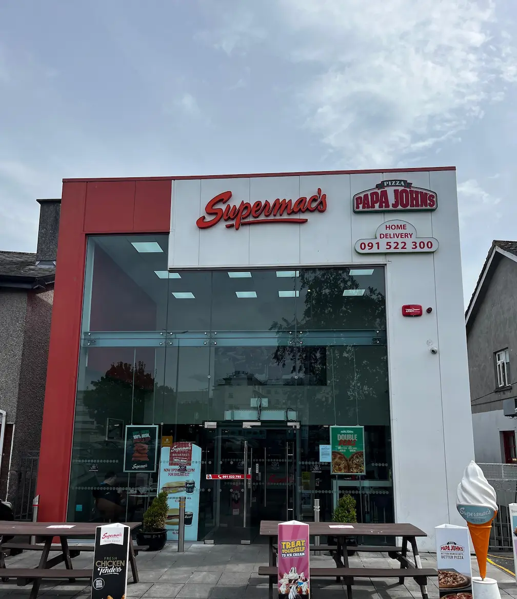 Supermac's and Papa John's restaurant exterior in Newcastle, Ireland. Modern building with red and white facade, large glass front, prominent dual branding, 