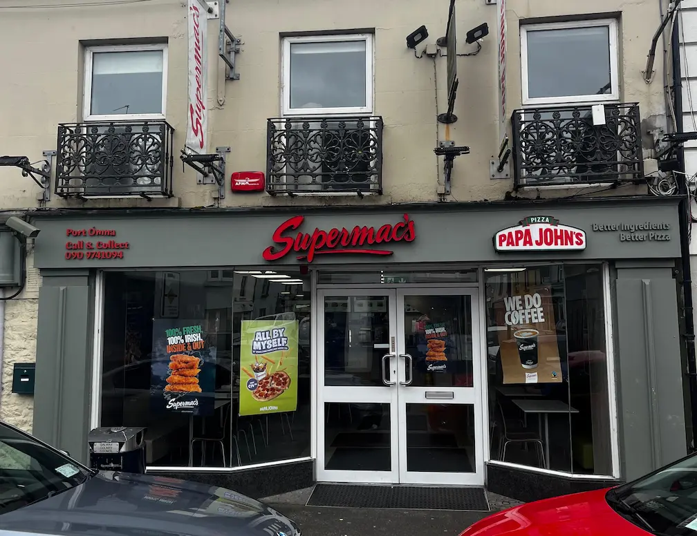 Supermac's and Papa John's restaurant exterior in Portumna, Ireland. Features prominent dual branding, a grey shopfront, and a traditional building facade with ornate Juliet balconies on upper windows.