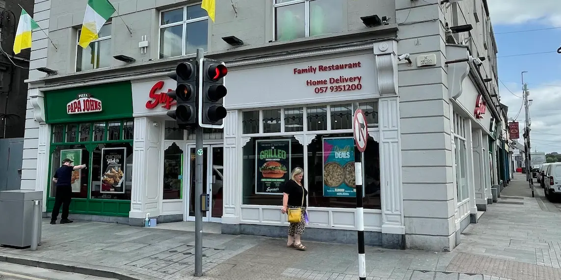 Supermac's and Papa John's fast-food restaurant exterior in Tullamore, Ireland. Offering takeaway tullamore, food delivery and pizza delivery in Tullamore and surrounding areas. Features prominent dual branding, traditional Irish building facade, green and white shopfronts, a traffic light, and Irish national flags.