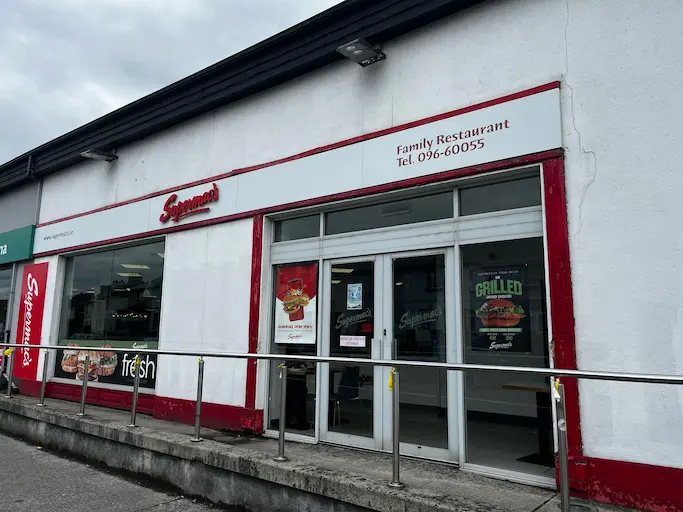 Supermac's Family Restaurant exterior at Casey's Service Station in Ballina, Ireland. Features a modern, single-story building with white walls, red trim, large windows, and