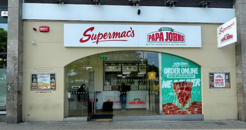 Supermac's and Papa John's restaurant exterior in Tallaght, Dublin, Ireland. Features prominent dual branding, a modern storefront with a large archway entrance, and a window displaying 