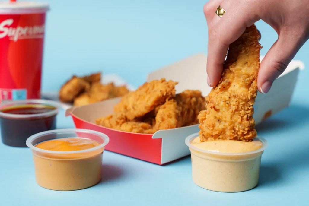 A Supermac's Chicken Tender being dipped into the new Secret Sauce with Korean BBQ and Smokey Honey dips alongside a box of Chicken Tenders
